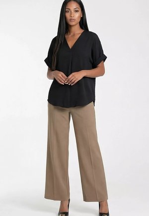 Kobieta stoi, ubrana w czarną krótką bluzkę z dekoltem w serek, beżowe szerokie spodnie i czarne szpilki, ręce splecione przed sobą na jednolitym tle.