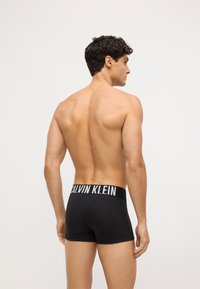 Czarne bawełniane bokserki z białym logo "CALVIN KLEIN" na gumce; model pokazany z tyłu, ustawiony lekko bokiem.