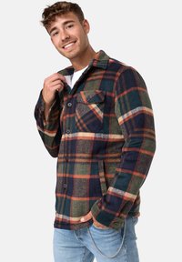 Camisa de franela a cuadros en verde, azul marino y naranja. Presenta un cuello, dos bolsillos delanteros y cierre de botones, fabricada en una suave tela texturizada.