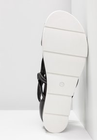 Sandale noire avec une semelle en caoutchouc blanche, dotée de détails en relief. Brides réglables en matériau doux. Étiquette de taille visible sur la semelle.