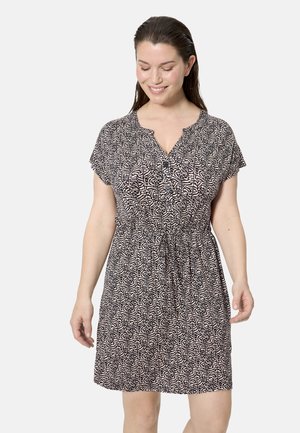 Mujer que lleva un vestido de manga corta con patrón en blanco y negro, con botones y cintura ajustable, sonriendo y mirando hacia abajo.