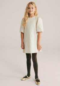 Svetlo kremna obleka s puff rokavi in teksturirano tkanino, kombinirana z črnimi bleščečimi nogavicami in zlatimi baletnimi copati. Preprost, eleganten dizajn.