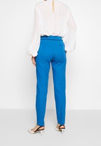 Een persoon draagt een vloeiende witte blouse en blauwe broeken, met chique hakken. De nadruk ligt op de levendige kleuren en de pasvorm van de outfit.