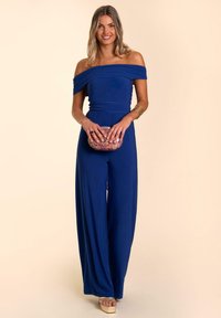 Mono azul marino ajustado, con escote off-the-shoulder y piernas anchas, hecho de una tela suave; modelo sosteniendo un clutch estampado, usando sandalias de plataforma.