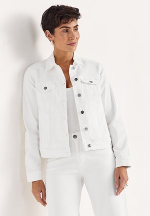 Frau mit kurzen dunklen Haaren trägt weiße Jeansjacke und Hose, steht vor einer hellen Wand und schaut zur Seite.