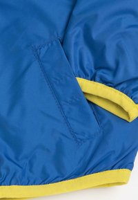 Veste bleue avec des bordures jaunes et une poche. Texture lisse, avec des bords surpiqués et un ourlet froncé pour un look ajusté.