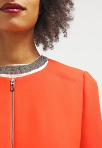 Oranžový blejzr se strukturovaným designem, s kovovým korálkovým límcem a předním zipovým uzávěrem, který zvýrazňuje hladkou texturu látky.