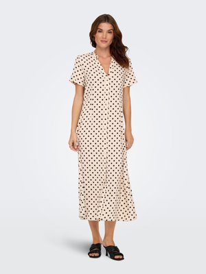 Femme debout portant une robe midi beige à manches courtes boutonnée devant, à pois noirs, et des sandales noires à talons.
