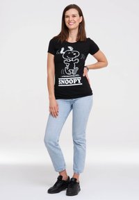 Camiseta negra con un gráfico de Snoopy en blanco y texto. Mangas cortas, escote redondo, combinada con jeans azul claro y zapatillas negras.
