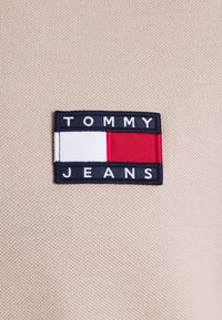 Beige tygbakgrund med en rektangulär marinblå ruta. Rutan har "TOMMY" i vit text, "JEANS" i vit text nedanför, samt röda och vita ränder.