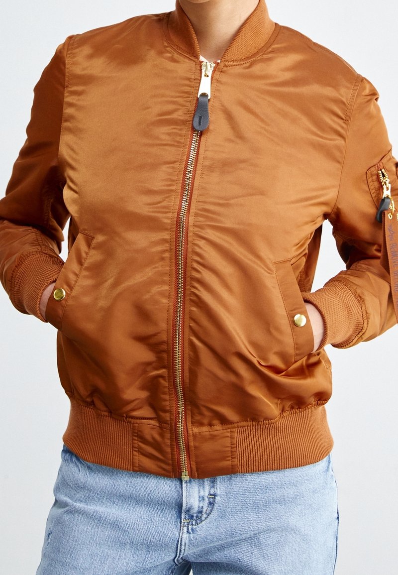 Personne portant une veste bomber couleur rouille avec poignets et taille côtelés, mains dans les poches, associée à un jean en denim bleu clair.