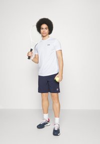 Camiseta atlética blanca de manga corta con el logo de FILA, pantalones cortos azul marino con acentos rojos y blancos, sosteniendo una raqueta de tenis y una pelota, usando zapatillas deportivas.