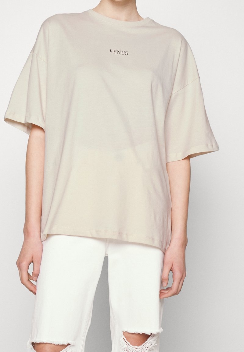 Camiseta de algodón color beige con mangas cortas y corte holgado, que presenta el texto "VENUS" en negro en el pecho. Combinada con vaqueros blancos rasgados.
