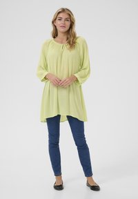 Blouse vert clair avec de longues manches légèrement bouffantes, détail de décolleté froncé, associée à un jean skinny bleu foncé et des ballerines noires.