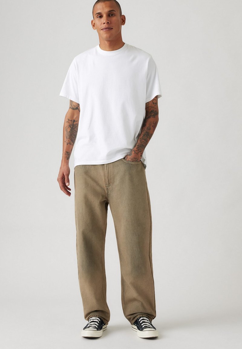 Camiseta blanca de algodón, corte relajado, combinada con pantalones de mezclilla caqui, de pierna recta, con una textura sutil y zapatillas negras de perfil bajo.
