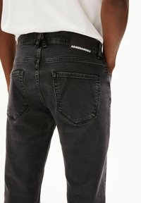 Achteraanzicht van een persoon die zwarte spijkerbroeken draagt met zakken en een merklabel met de tekst "ARMEDANGELS" op de tailleband.