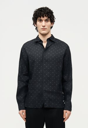 Άνδρας με σγουρά σκούρα μαλλιά, που φοράει μαύρο πουκάμισο με μικρά λευκά σχέδια ιστιοφόρων, στέκεται μπροστά σε απλό ανοιχτό φόντο.