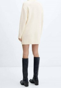 Osoba v dlouhém smetanovém svetru a kožených botách po kolena, stojí proti jednoduchému světlému pozadí, otočená zády.