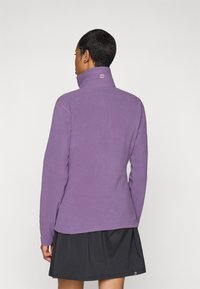 Lila Fleece-Pullover mit Stehkragen, langen Ärmeln, kontrastierenden Nähten und einem Markenlogo am hinteren Kragen. Über einem schwarzen Rock getragen.