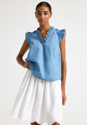 Kvinde iført ærmeløs blå denimbluse med flæsekant og hvid midi-nederdel med broderi, stående foran en ensfarvet baggrund.