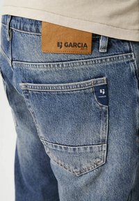 Jeans de denim en azul claro con una textura desgastada. Incluye una etiqueta de cuero con "GARCIA" en la parte trasera y una etiqueta de tela con la marca en el bolsillo.