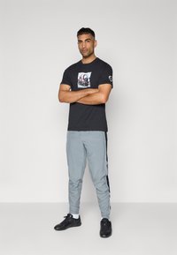 Sort T-shirt med grafisk design, grå træningsbukser med sorte sidelænder og sorte sportssko med hvide detaljer.