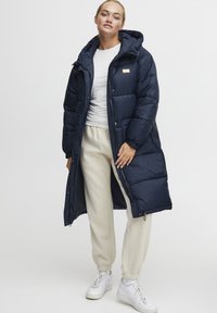 Navy blue padded long coat with hood, featuring a front zipper and snap buttons, worn over a white shirt and beige joggers.