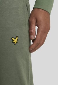 Grüne Sweatpants aus strukturiertem Stoff, mit einem gelben Vogel-Logo auf dem linken Oberschenkel. Die Hand befindet sich in der Nähe der Seitenaht.