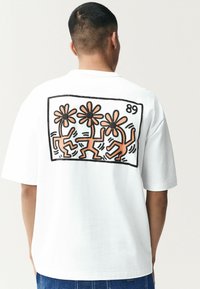 Balta trumpo rankovio marškinėlių su spalvinga grafika, vaizduojančia tris šokančius figūras ir gėles ant nugaros, turinčiosiomis storas juodas kontūras.