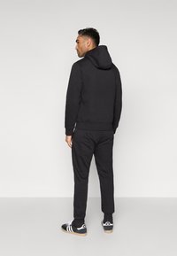 Black hooded sweatshirt with a textured finish, paired with matching joggers. Model stands with back facing, wearing black and white sneakers.