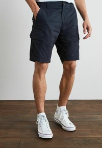 Marinefarbene Cargo-Shorts mit Seitentaschen, aus leichtem Material gefertigt. Verfügt über einen klassischen Schnitt und ein Design mit mehreren Taschen. Getragen mit weißen Sneakers.
