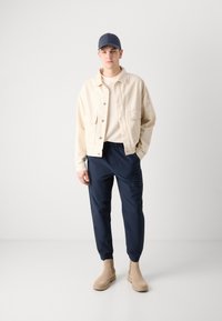 Beige denim jack, marineblauwe trainingsbroek, beige enkellaarzen en een marineblauwe pet. Eenvoudig ontwerp met een losse pasvorm en minimale hardwaredetails.
