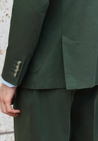 Dunkelgrüne maßgeschneiderte Sakko mit einem eleganten Design, subtiler Textur, vier dekorativen Knöpfen am Ärmel und einer Seitentasche.