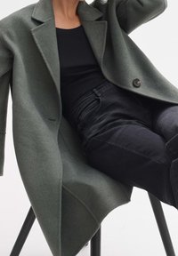 Personne portant un manteau en laine vert foncé sur un haut noir et un pantalon noir, assise sur une chaise noire devant un fond blanc.