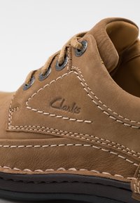 Chaussure en cuir marron avec des détails cousus, des œillets métalliques et des lacets beige. Présente une surface texturée et un logo "Clarks" embossé.