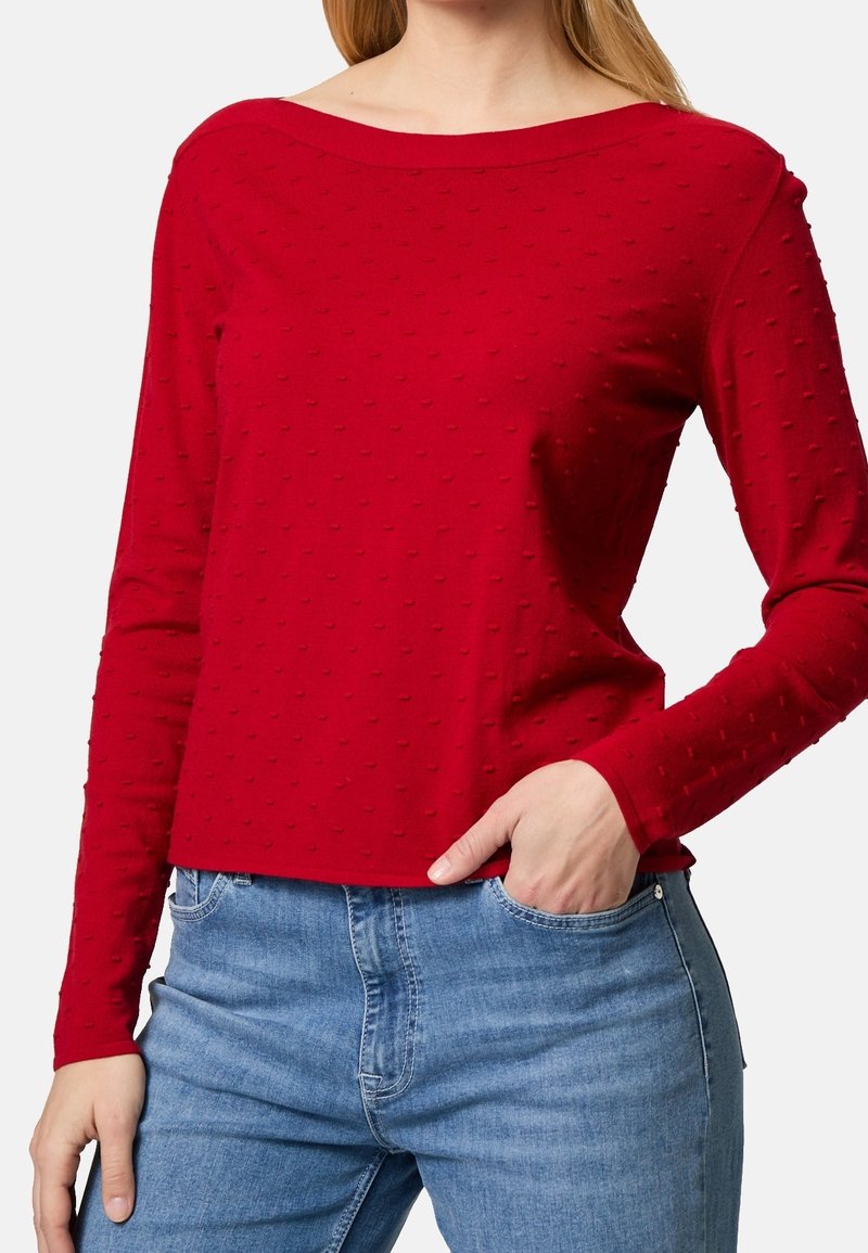 Pull en tricot rouge à manches longues, présentant un motif texturé avec de petits points en relief, un col large et des manches ajustées, associé à un jean bleu clair.