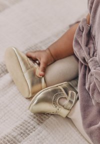 Chaussures bébé en cuir doré avec un bout arrondi et un design découpé, dotées d'une bride et d'une semelle souple en daim, posées sur une couverture texturée.