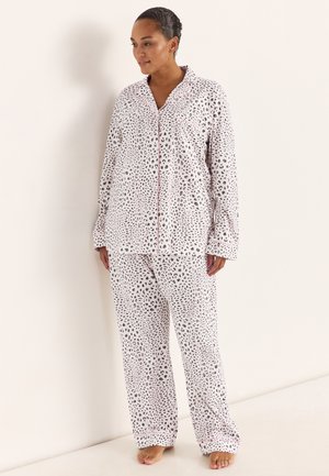 Frau in weißen und grauen Leopardenmuster-Pyjamas mit pinkfarbenen Paspeln, steht barfuß vor einer schlichten hellen Wand.