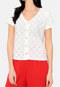 Blusa bianca a maniche corte con motivo in pizzo, scollo a V e chiusura con bottoni. Tessuto strutturato, orlo ondulato discreto, abbinata a pantaloni rossi.