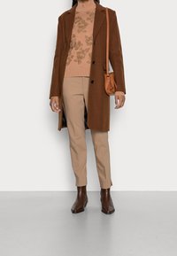Manteau en laine marron par-dessus un pull beige à motif floral, pantalon beige et bottines marron. Comprend un petit sac marron.