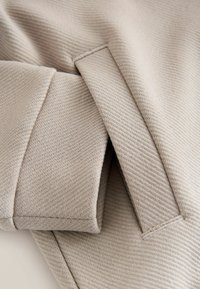 Beiger, strukturierter Stoff mit diagonalem Muster, einer gefalteten Ärmelpartie und geraden Ziernähten für ein maßgeschneidertes Erscheinungsbild.
