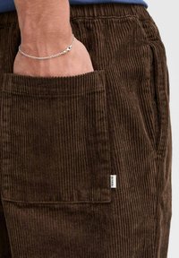 Braune Cordhosen mit vertikalen Rippungen, ausgestattet mit einer Seitentasche und einem kleinen Logotag. Die knitterfreie Textur verstärkt die visuelle Tiefe.
