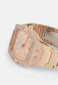 Quadratische roségoldene Uhr mit Kristallakzenten, ausgestattet mit einem geometrischen Zifferblatt und drei Teilzifferblättern. Metallarmband mit glänzender Oberfläche.