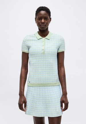 Vrouw draagt een korte mouw, groen-blauw geruit polohemd met bijpassende rok, staand tegen een effen witte achtergrond.