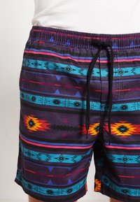 Pantalones de natación con un patrón geométrico colorido. Tienen una base púrpura, cinturilla elástica con cordón y acentos llamativos en rojo y azul.
