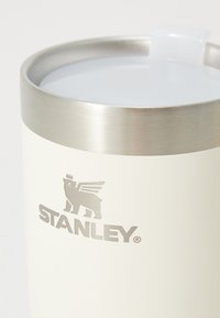 Baltas metalinis puodelis su nerūdijančio plieno kraštu ir logotipu, vaizduojančiu liūtą su sparnais. Dangtelis turi skaidrų plastikinį atidarymą gėrimo siurbimui.