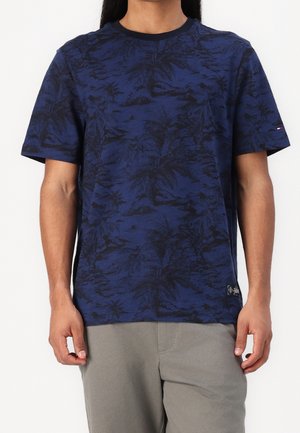 Mann trägt marineblaue kurzärmelige Tropen-T-Shirt mit Muster und hellgraue Hose, steht vor einem einfarbigen Hintergrund.