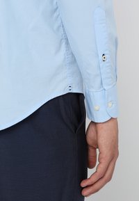 Camisa de algodón azul claro con cuello puntiagudo, puños abotonados y dobladillo curvado, que presenta sutiles detalles de costura y una textura suave.