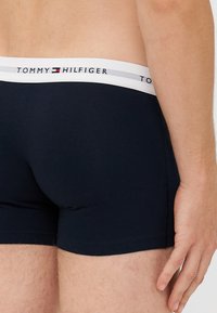 Sötétkék pamut boxeralsó fehér elasztikus derékkal, melyen ismétlődő logó és szürke díszcsík található. Simás textúra, testhezálló fazon.