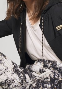 Svart zip-up jacka med texturerad tyg och en logotyp med guldaccenter, tillsammans med en ljusfärgad skjorta och mönstrade byxor i svart, vitt och guld.